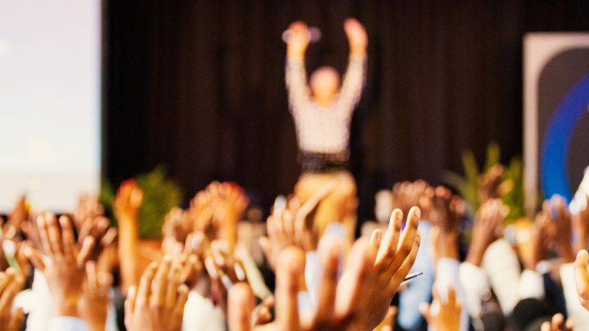 An image focusing on the celebrating audience's hands with a blurry man raising his hands on stage in the background.