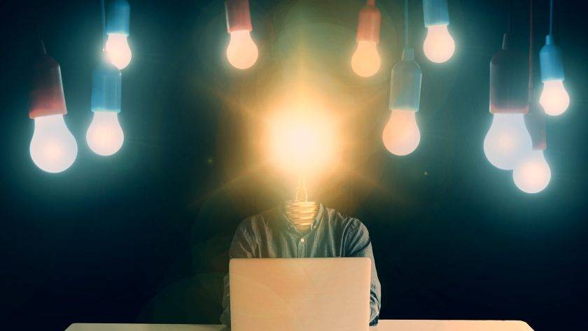 An image of a person with a lit up lightbulb in place of the head. The person is on a computer and has lit up lightbulbs hanging over him in the background as well.